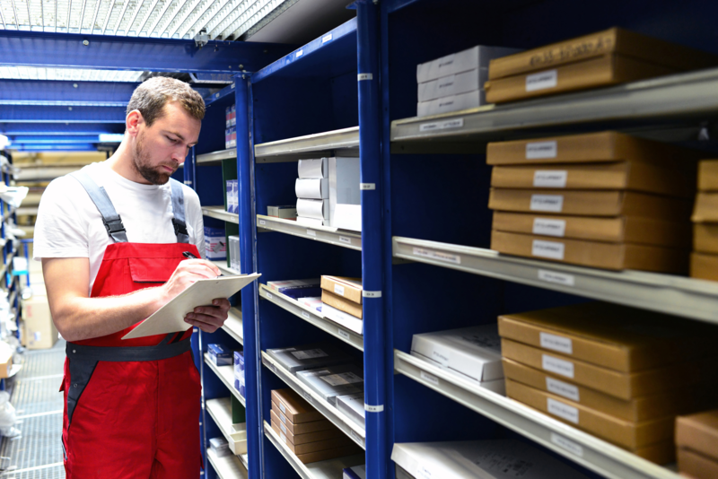 A worker uses a clipboard to check spare parts inventory in a store room stocked with boxes.